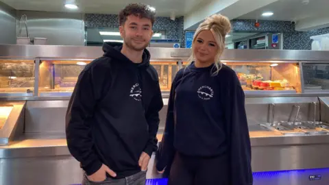 Mitch and Destiny inside Merchants chip shop. They are in front of a counter with lots of fried food and smiling for the camera. 