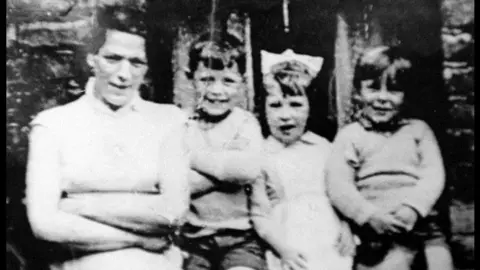 PA Media A black and white photo of a woman standing looking at the camera with her arms folded. Beside her are three children, also looking at the camera and smiling.