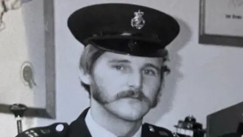 Family Handout A black and white image of a male police officer in uniform. He has a thick, wide moustache leading into sideburns. He is wearing a black peaked police cap with a badge in the centre. A white collar and black jacket can be seen, with  shoulder epaulettes visible, He is standing in front of a white wall with undiscernible office equipment behind.