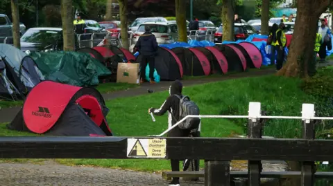 PA Media An early morning operation to remove tents which have been pitched by asylum seekers along a stretch of the Grand Canal, Dublin. There are numerous tents and people in high-viz jackets and a garda can be seen in the background.