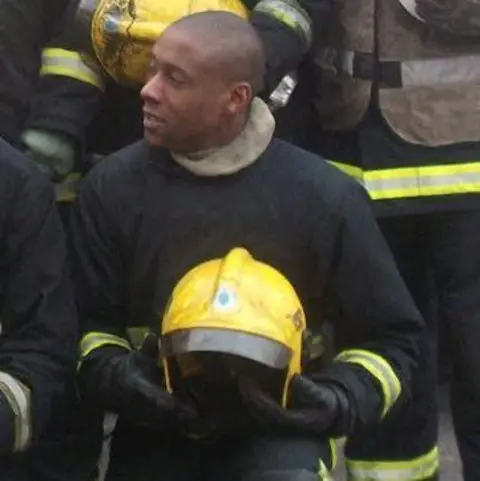 Jez Daniels Jez Daniels in his firefighter's uniform, holding a yellow helmet and with colleagues behind him