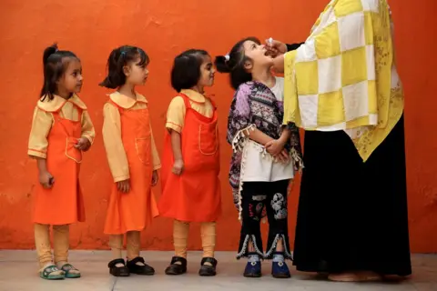 BILAWAL ARBAB/EPA Three children in red pinafore dress uniforms queue behind a child in a black and purple shawl as she receives an oral vaccination in Peshawar, Pakistan, 28 October 2024.  An adult wearing a black dress and a yellow and white check shawl is putting the droplets into the child's mouth.