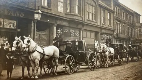 Eddie Wulff Pictured here is the wedding procession of Max and Mabel Wulff. Six large black and white horses pull three carriages along Alexandra Road in Pill, South Wales. The photograph is black and white and depicts a typical high street in the UK at the turn of the century.