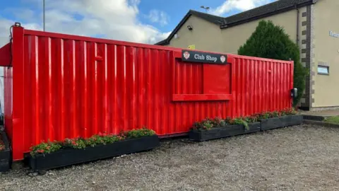 A large red metal shipping container. It has a close hatch in the middle and sign saying "club shop". In front a black planters with flowers.