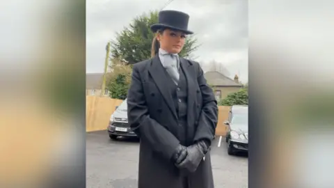 Hollie James A young woman stood in black suit and black top hat on with black hearses in the background