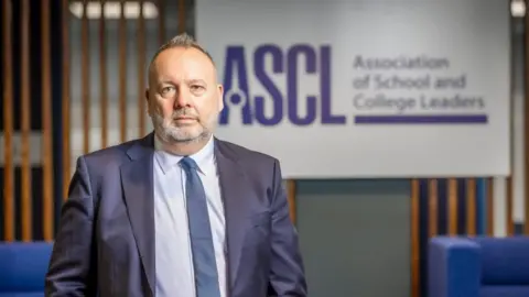 Association of School and College Leaders Pepe Di'Iasio, wearing a blue suit, blue tie, and and a light blue shirt. He has a grey beard, receding hair which is light brown and is spiked at the front. He is standing near a wooden screen, with a board stating ASCL, Association of School and College Leaders, and nearby are some blue sofas.