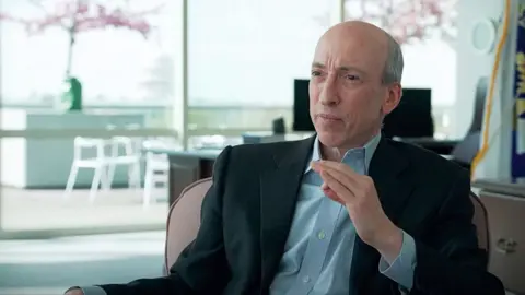 Gary Gensler, head of the US Securities and Exchange Commission, sits in an office, gesturing with his hand. He is bald, and heis wearing a dark jacket suit and open-necked blue shirt.