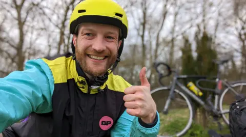 James Dunn  James is crouching in front of the camera and has his thumbs up. He is wearing a bright yellow bike helmet and a multicoloured waterproof jacket. 
In the background, out of focus you can see his black bike. 