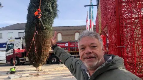 John Devine/BBC Paul Hicks has short grey hair with a grey beard and moustache and appears to be using his right arm to keep the leaning tree upright. He is wearing a grey overcoat with a white shirt underneath, with the collar up. Behind him is the leaning tree, with netting on it and a man in an orange hi-vis jacket and hard hat pulling the netting off