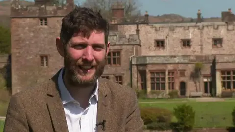 Ewan Frost-Pennington, who has a beard and is wearing a brown tweed jacket, pictured outside Muncaster Castle where he houses staff to keep it running