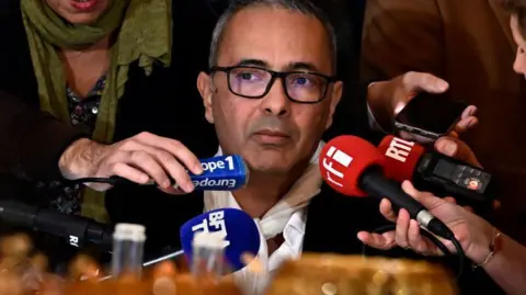 Getty Images Kamel Daoud looks on pensively as he answers questions from a throng of journalists. He is sat at a table and surrounded by hands holding microphones, audio recorders and smartphones.