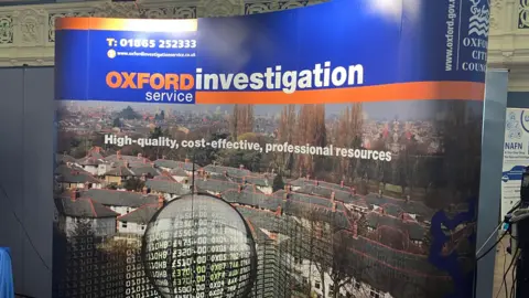 A sign at Oxford's annual Fraud Conference saying "Oxford Investigation Service". The tagline underneath reads "High-quality, cost-effective, professional resources". The sign shows a drone picture of housing, with a magnifying glass superimposed on front. Within the magnifying glass there is a list of monetary amounts and postcodes.