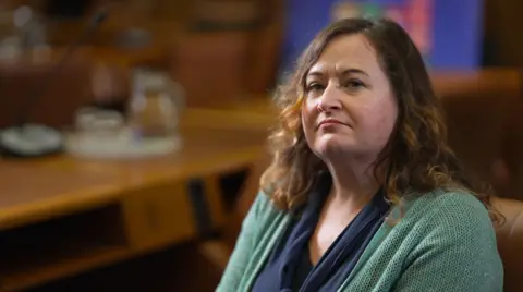 Claire Healy of the UN’s Office on Drugs and Crime (UNODC), sitting in an office looking at the camera, with a desk behind her. She is wearing a green cardigan and a blue top underneath. Her brown hair with blonde highlights is untied.