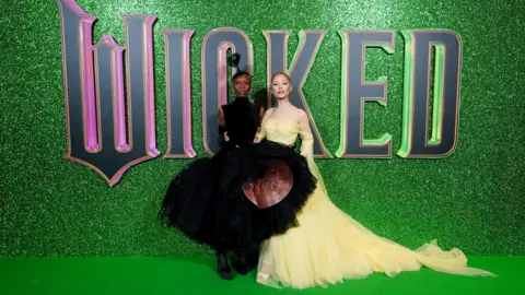 PA Media Cynthia Erivo and Arianna Grande pose together wear flowing black and yellow ball dresses in front of large Wicked lettering, during the premier event for the movie in the London