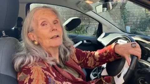 Sally has long grey hair and a red patterned dress and is at the wheel of her car