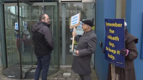 A cinema-goer walks past two protestors on his way into the cinema. The protestors are holding placards. One of the placards reads in yellow writing on a blue background: "Remember the Sabbath day, to keep it holy."