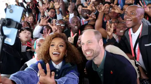 Getty Images Prince William and Nomzamo Mbatha pose for a selfie with young people during the Earthshot Prize Climate Leaders Youth Programme in Cape Town, South Africa. Both are smiling and are surrounded by dozens of smiling young people.