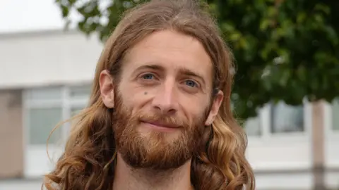 Josh has shoulder length brown wavy hair and a full beard. He is wearing a black T-Shirt. He is standing in the street and in the background you can see a block of flats and trees. 