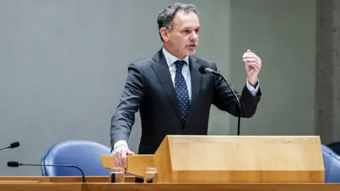 EPA-EFE/REX/Shutterstock Dutch foreign minister Caspar Veldkamp gestures with his hand as he speaks at a wooden lectern in the Hague. He wears a grey suit, a white polka dot navy tie and white shirt and has short dark grey hair