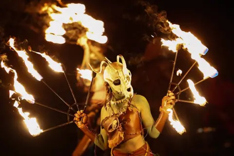 Jeff J Mitchell/Getty Images A costumed woman wearing a mask performs a fire-dance. She is painted green and holds a metal metal frame of five flaming torches in each hand