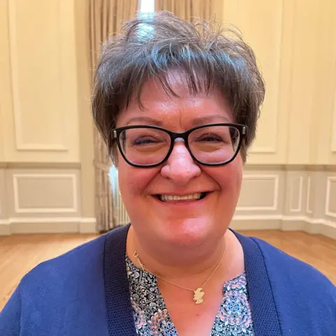 Dr Christiana Bratiotis beams at the camera while wearing a ditsy print top and a royal blue cardigan. She is also wearing a gold Scotland map necklace. She is standing in a grand room with high ceiling, a big window with fancy swap curtains and wall mouldings