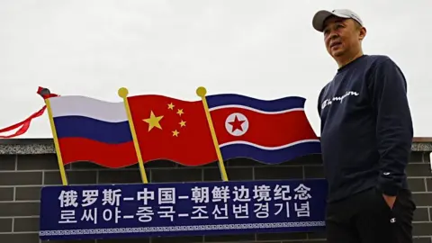 A man in a cap stands with his hands in pockets in front of cutout showing a row of three flags - Russia, China and North Korea. 

Below the flags, the three countries' names are spelled out in Mandarin and Korean. 