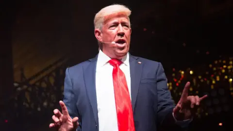 Mike Osman Mike Osman dressed as Donald Trump. He is wearing a blonde wig, a blue suit and red tie.