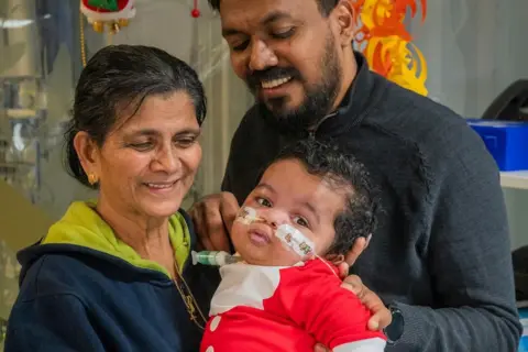 Teena Thomas Gabriel's granny is wearing a blue hooded top and has black hair tied up and gold earrings. She is looking at her grandson while holing him in her arms and smiling. Gabriel is wearing a red Santa outfit and has tubes in both nostrils that are Sellotaped to his face. His dad is in the background in a dark top and smiling while looking at his son.