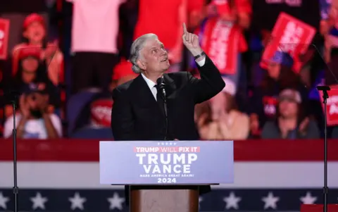 Reuters Rev Franklin Graham points upwards whilst standing in front of a Trump Will Fix It stand at a rally