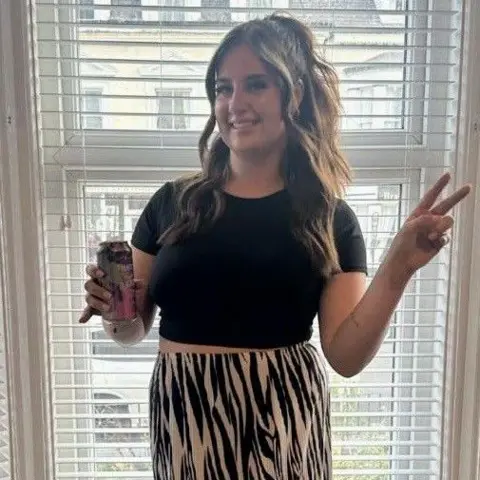 Paige Roberts Paige Roberts standing in bay window wearing zebra print trousers and a black belly top. She is smiling and holding a can in one hand and throwing up the peace sign with her other 