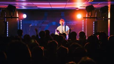 Ella Calver A concert sees man with guitar singing to crowd. It is dark and only silhouette of heads care seen. 