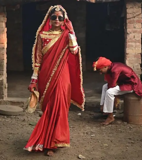 Innovation for Change A girl models at a fashion show that has gone viral, she is walking down a street while wearing colourful red clothing, jewellery and sunglasses, and a man is sitting on the side of the street behind her putting his shoes on.