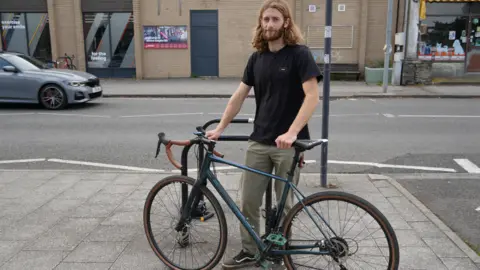 Josh is standing on the pavement with his bike in front of him. 