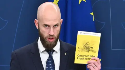 Getty Images Sweden's Minister for Civil Defence Carl-Oskar Bohlin holds a copy of  the new version of the preparedness booklet "If the crisis or war comes". The pamphlet has a yellow cover and an image of a woman in army fatigues holding a large gun.