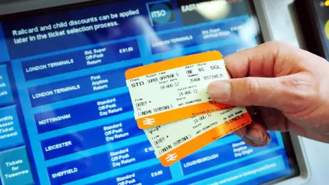 PA Media Two kinds of rail tickets are held up in front of a ticket machine displaying a variety of destinations in the UK