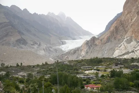 The Passu glacier can be seen in the distance, running between two mountains, with a village and dense tree cover in the foreground.