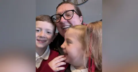 Debbie McCrudden Debbie McCrudden, a woman with dark hair, a nose ring and black-framed glasses, holding her two children on her lap. Everyone in the photo is laughing.