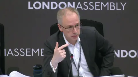 London Assembly Richard Watts sits in front of a backdrop that says London Assembly. He is wearing a dark suit jacket and a white open-necked shirt, as well as a lanyard. There is a slim microphone on the table in front of him. 