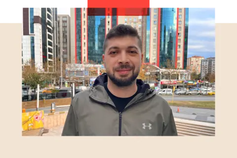 Photo of Hussam - a young adult man with a moustache and trimmed beard. He is staring straight to camera. Behind him is a road and some high rise buildings.