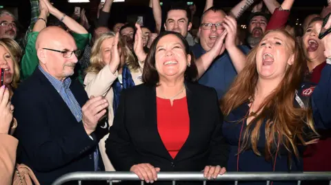 Getty Images Mary Lou McDonald celebrates with her supporters after being elected at the RDS Count centre in 2020. She has a wide smile and she's surrounded by people cheering and clapping.