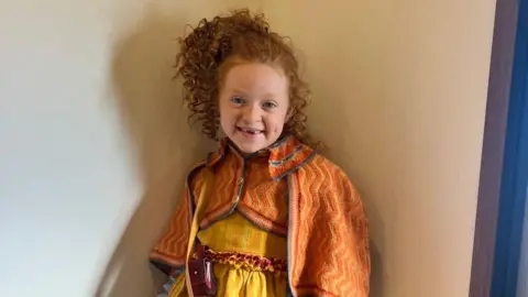 Lauren Kelly A young girl with curly, ginger hair is wearing bright clothing whilst smiling at the camera.