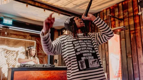 Getty Images Jordan Adetunji performing onstage. He's wearing a stripped top and black trousers and holding the mic over his head. A dj can be seen behind him.