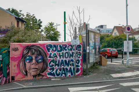 Getty Images A mural near a pedestrian crossing, with a picture of a woman with a short bob and round sunglasses with the words: "Shame must change sides"