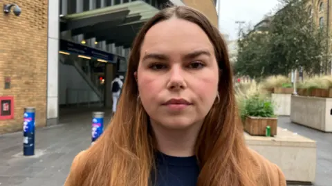 A close up of Molly, who is looking into the camera with a straight face. She is stood outside a train station. She has long brown hair and is wearing a dark blue top with small, hooped earrings. 