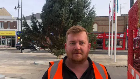 John Devine/BBC Daniel Fortuna has red hair and a beard with a moustache. He has a black T-shirt and a hi-vis orange tabard. Behind him is the bottom section of the Christmas tree, which is leaning slightly to the right. There are flagpoles and part of the town's war memorial on the right, with shops in the background