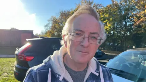 Mike Thurland with glasses and grey hair stands in a car park in front of two cars.