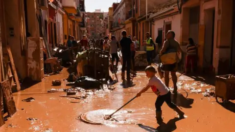 Reuters People seen sweeping muddy floodwater from a street in Valencia. Damaged furniture can also be seen piled up. 