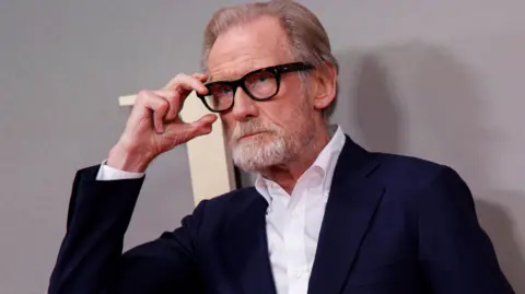 EPA Actor Bill Nighy arrives for the premiere of the film 'Joy' during the BFI London Film Festival at Royal Festival Hall. He is raising his right hand to touch the frames of his black spectacles as he poses in front of the cameras on the red carpet.