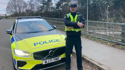 BBC A police officer wearing a high viz vest standing in front of a police car