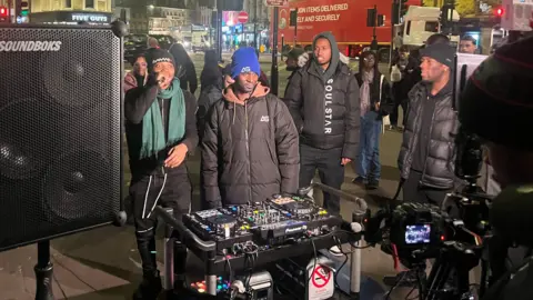 BBC DJ decks on a trolley with a big speaker on the left. There's a camera in front of the DJ as he goes live on Tik Tok. Behind the decks, there's a man with a blue had, brown hoodie and black puffer jacket with the logo AG on it. He's flanked by a man in a gree scarf rapping  as well as onlookers. 